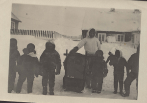 Image of Teacher by backpack, and six school children. One girl has doll. 