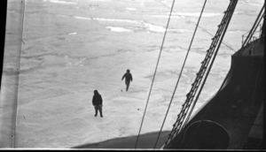 Image of Two men on ice near ship; teams in distance