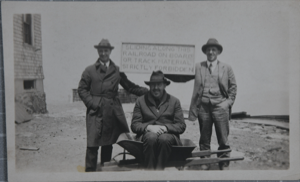 Image of Donald MacMillan (rt) with two unidentified men, holding a sign saying "Sliding along this railroad on board or track material strictly forbidden"