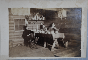 Image: Interior of a log cabin with men at a dining table.