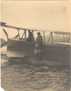 Image of Donald MacMillan in furs, on navy plane pontoon. Pilot in cockpit