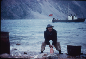 Image of Inuit man with trout. Fishing vessel beyond.