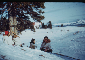 Image: Inuit women by evergreen tree in a snow covered yard.