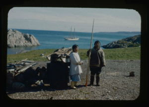Image of Eskimo [Inuit] couple by sledge and barrels. He holds narwhal tooth. BOWDOIN in distance