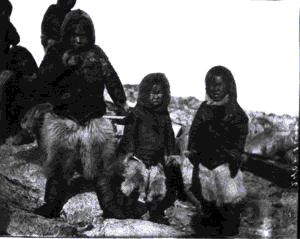 Image of 3 young Inuit boys in polar bear pants. Adults beyond.