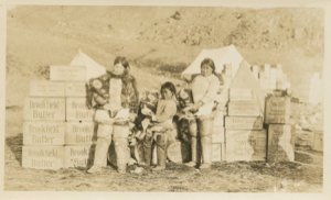 Image of Ane Petersen, Inuit girl and woman by crates of supplies. Each is holding a Swi