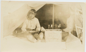 Image of Inuit man sittiing at tent opening, holding pan of food. Snowshoes at right