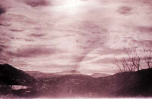 Image: Clouds, distant mountains seen thru break in near-mountains; bare decidious trees on the right