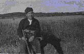 Image of ? seated in field with gun. Raven [dog] beside him