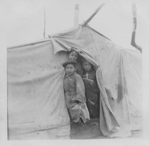 Image of Three Indian [Innu] children in tent doorway. One wears large cross