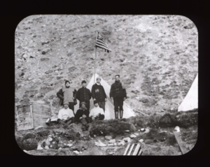 Image: Seven expedition members by tent and pole with U.S. flag