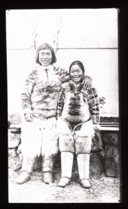 Image of Inuit couple standing by building, rifles on wall