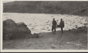 Image of Two expedition men carrying large rock on stretcher. Ice pack beyond