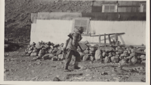 Image of Expedition man with rifle walks by Borup Lodge. Note foundation
