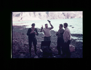 Image of Four crewmen on beach with beer (?) bottles. One has camera  [purple] 