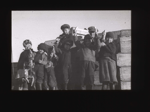 Image: Group of boys by wooden crates; each holds up an banana  [b&w]