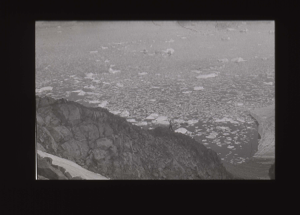 Image of Looking down to crewman on rocks, waving. Ice beyond. Glacier in foregroun [b&w]