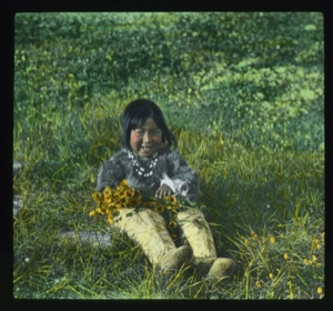 Image: Shoo-e-ging-wa [Suakannguaq Qaerngaaq] sitting on grass holding flowers