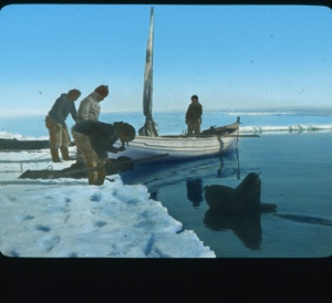 Image: Small sail boat by shore; three men, Inuit boy on stomach, woman ....