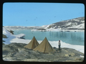 Image of Two tents just above ice foot. Inuit man with pipe standing by