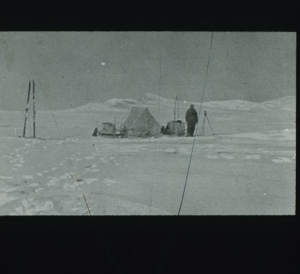 Image of Long view to tent site. Man stands beside it. Tall tripod, sledge with skis 