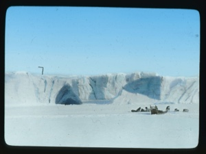 Image: Dog team and sledge near glacier face which has large cave-like opening