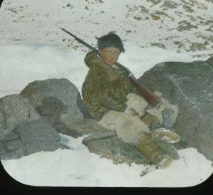 Image of Inuit boy sitting among rocks with rifle over his shoulder....