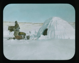 Image of Inuit man with pipe outside snow igloo.Equipment near