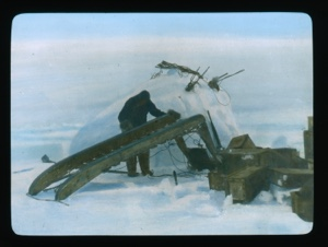 Image: Man by snow igloo working on runners of inverted sledge. Wooden crates near