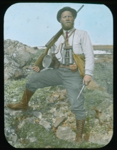 Image of Elmer Ekblaw standing on rocks with rifle, knife, binoculars, canteen