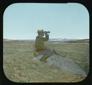 Image of Team member sitting on tundra wearing straw hat, using binoculars