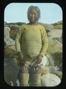 Image: Old Esquimeau woman by stone igloo, holding piece of hide