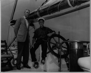 Image: Donald MacMillan seated at the BOWDOIN's wheel. ? standing by, after arriving in Mystic