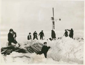 Image of Six men by windsock pole. Two others shovel snow off their tent. Soviet Polar Expedition