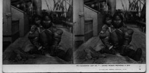 Image of Eskimo [Inuk] woman [with toddler on her back] preparing a skin [aboard ship]