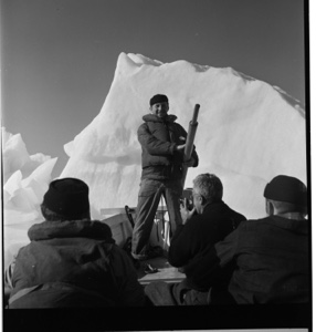 Image of Joe Mattiacu standing in bow of boat holdiing oar [?] Men seated. Iceberg behind.