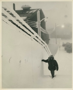 Image: Man walking up incline by wall; snow-encrusted railings, street lamp and overhead wires