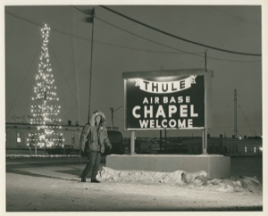 Image of Man near Base Chapel sign. Christmas "tree" lights near. Thule AFB