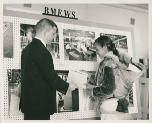 Image of RCA employee presents papers to Inughuit woman by BMEWS display board [Aqattaq Qujakitsoq and baby Atangana]