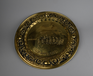 Image: decorative brass platter with embossed design of Umaanaq, man with dog sledge, and word 