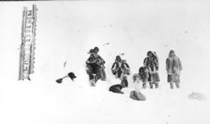 Image of Inuit and dogs in snow; up-ended sledge nearby