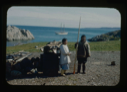 Image of Eskimo [Inuit] couple by sledge and barrels. He holds narwhal tooth. BOWDOIN in distance