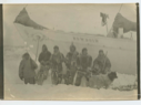 Image of Crew members in furs, beside iced-in Bowdoin; some with snowshoes. Dog