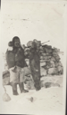 Image of Inuit man with seal in front of stone cache. Harpoon near