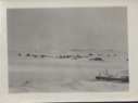 Image of Village in winter. Vessel iced in, in harbor. Fishing boom(?) in foreground