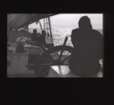 Image of Crewman at  wheel, others sitting beyond  [b&w]