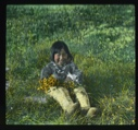 Image of Shoo-e-ging-wa [Suakannguaq Qaerngaaq] sitting on grass holding flowers