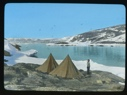 Image of Two tents just above ice foot. Inuit man with pipe standing by