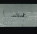 Image of Long view to tent site. Man stands beside it. Tall tripod, sledge with skis 