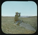Image of Team member sitting on tundra wearing straw hat, using binoculars